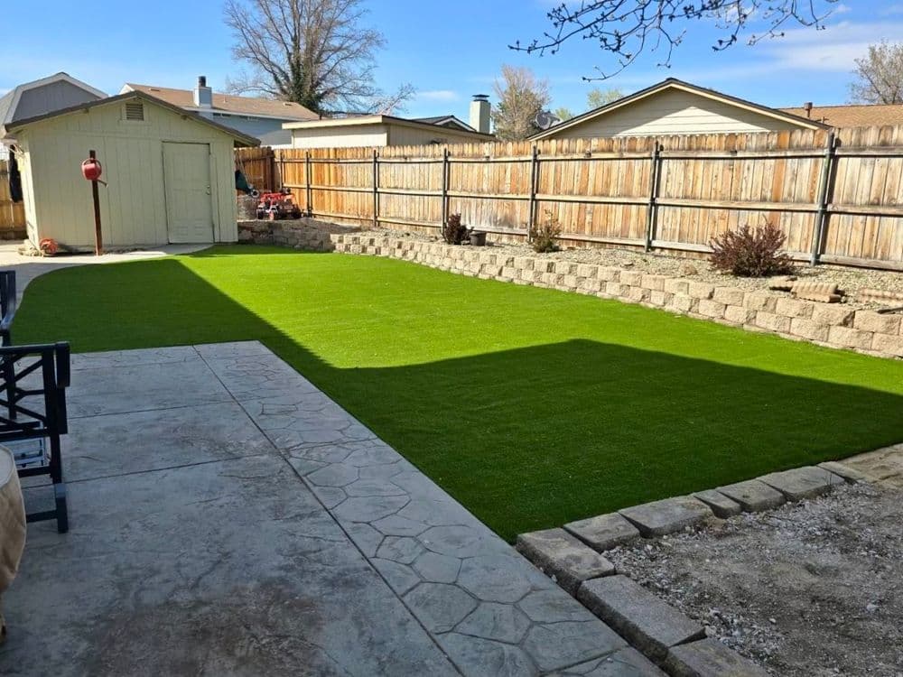 Lush green artificial grass lawn in a backyard with stone borders and wooden fence.