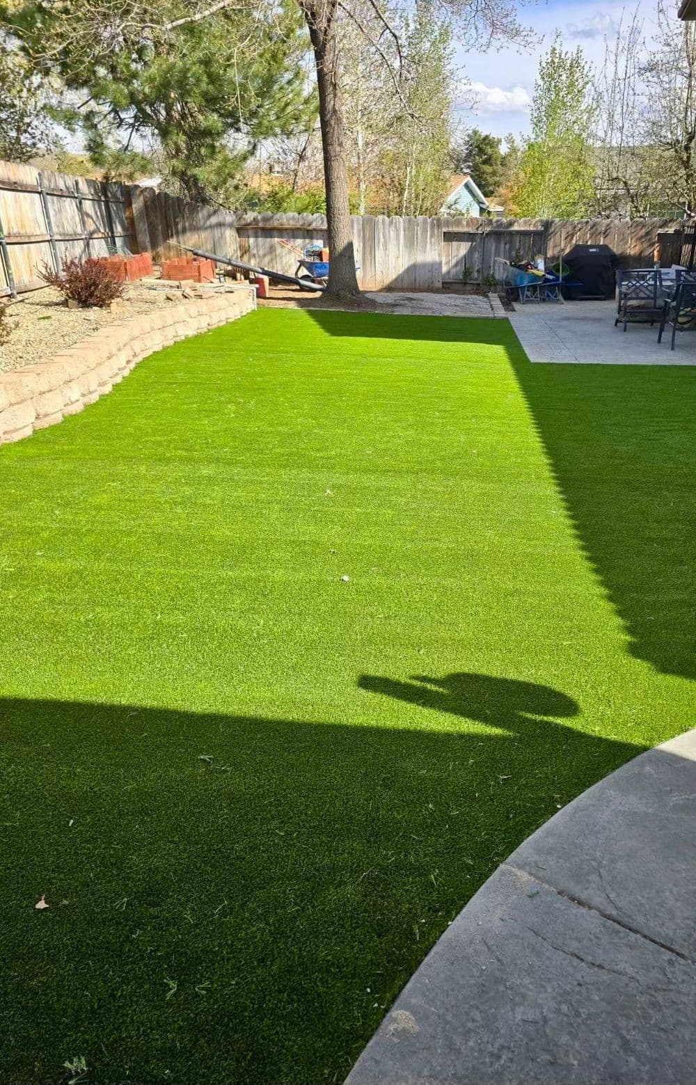Lush green artificial grass lawn in a sunny backyard setting with trees and patio.